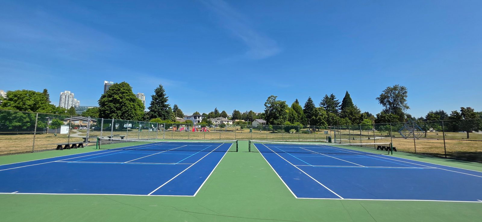 Empty blue pickleball and tennis courts at Ernie Winch Park, with highrises in the distance.