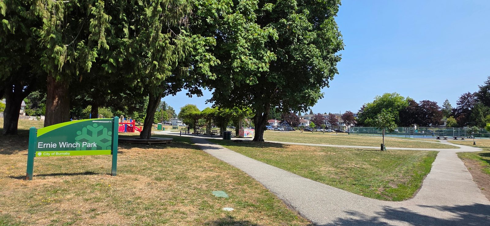 Ernie Winch Park entrance sign on a sunny day, with pathways, trees and courts beyond.